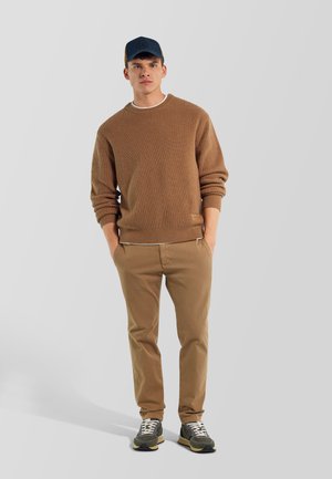 Giovane uomo che indossa un maglione marrone lavorato a maglia, pantaloni beige, scarpe da ginnastica scure e un berretto da baseball blu navy, in piedi con le mani in tasca.