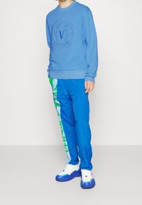 Sweatshirt bleu avec un logo en relief, associé à un pantalon bleu avec une bande graphique verte. Des baskets blanches et bleues complètent le look.