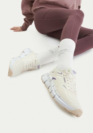 Person sitting on floor wearing mauve leggings and white Reebok sneakers with thick soles and white socks with logos.
