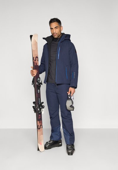 Tamsiai mėlyna slidinėjimo striukė su mėlynomis detalėmis, juodas vidurinis sluoksnis ir derančios kelnės. Asmuo laiko slides ir slidinėjimo akinius, dėvi tamsius batus ir įrangą.