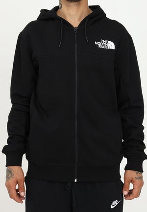 Mies, jolla on musta vetoketjullinen huppari, jossa on The North Face -logo rinnassa, ja mustat Nike-housut. Kädet näkyvissä, ranteissa tatuointeja.