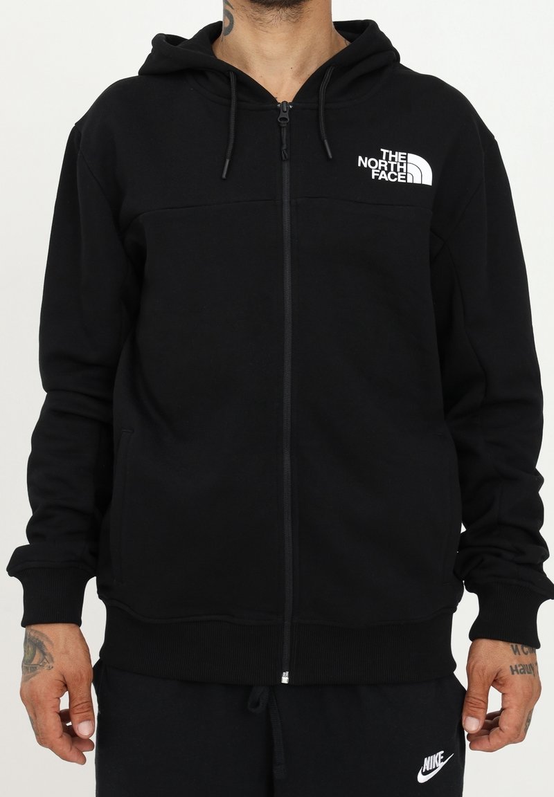 Homme portant un sweat à capuche noir zippé avec le logo The North Face sur la poitrine et un pantalon Nike noir, mains visibles avec des tatouages sur les poignets.