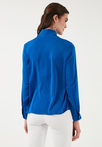 Cămașă cu mânecă lungă de culoare albastră strălucitoare, cu o textură netedă și un guler clasic. Vederea din spate arată fante laterale și detalii cu nasturi pe mâneci.