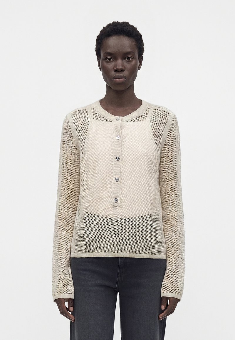 Femme aux cheveux courts portant un haut beige en maille à manches longues boutonné et un pantalon foncé, debout devant un fond blanc uni.