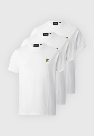 Homme portant des lunettes de soleil, un t-shirt blanc avec un petit logo jaune, un pantalon noir, des baskets rouges et un collier de perles, debout avec une main dans la poche.
