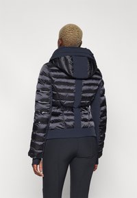 Tumed puffer-jakk on riidekohtadega, kapuuts, elastsed mansetid ja kontrastselt matt seljaosa. Sile, läikiv tekstuur koos funktsionaalsete detailidega.