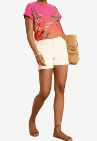 Różowa koszula z krótkim rękawem w kwiatowy wzór, sparowana z beżowymi szortami, uzupełniona plecioną torbą i brązowymi sandałami.