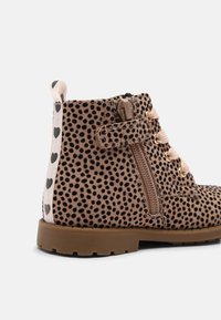 Bottine en daim marron avec des pois noirs, lacets beige, fermeture éclair latérale, bande velcro, et languette blanche avec des imprimés de cœurs noirs.