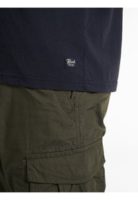 Ourlet de chemise bleu marine avec logo Petrol Industries au-dessus de la poche d'un pantalon cargo vert olive sur fond blanc.