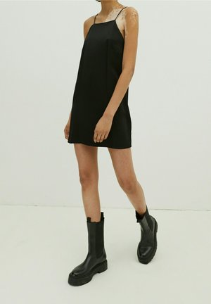 Vestito nero senza maniche con taglio dritto e spalline sottili, abbinato a stivali neri in pelle al ginocchio con suola spessa.