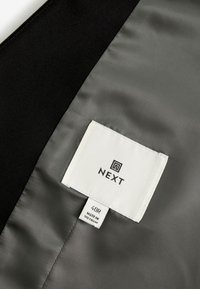 Clothing label inside a garment showing brand "NEXT," size "40R," and "Made in Vietnam" on smooth gray and black fabric.