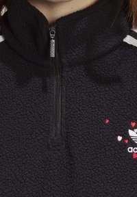 Svart fleece-pullover med hög krage, halvdragkedja fram och broderad logotyp. Har färgglada hjärtaccenter runt logotypen.