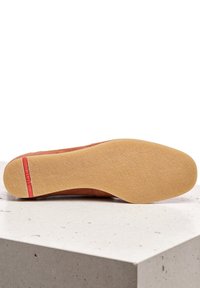 Brązowy zamszowy but wsuwany z teksturowaną gumową podeszwą, z czerwonym akcentem na pięcie. Podeszwa jest płaska i elastyczna.