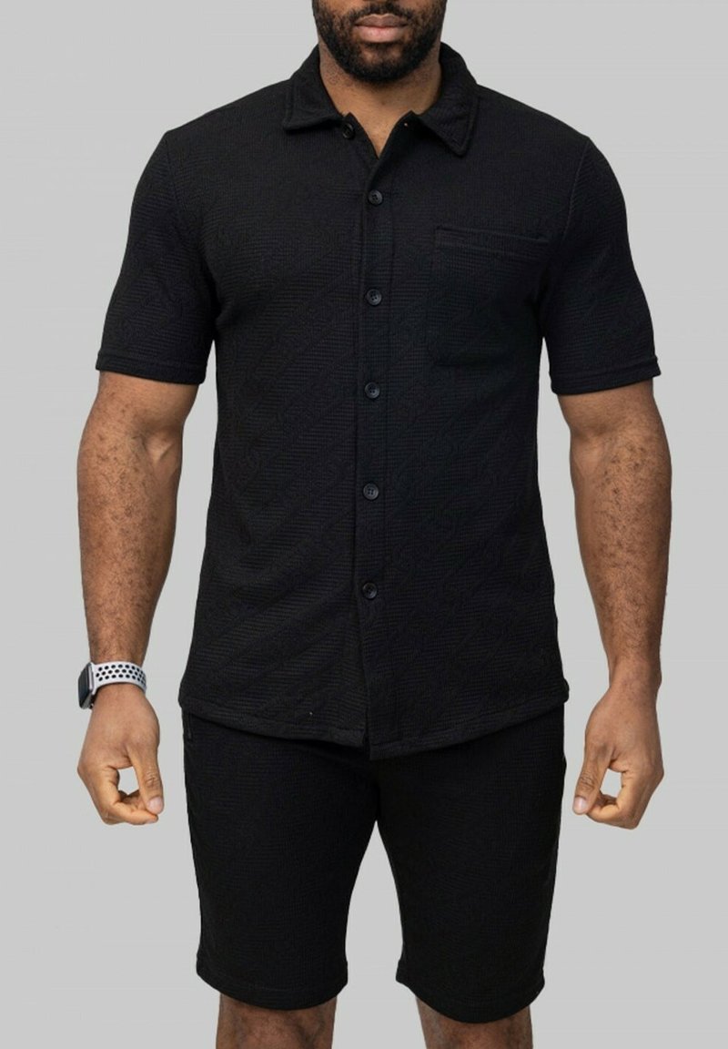 Chemise noire à manches courtes avec un motif texturé, des boutons sur le devant et une poche sur la poitrine, associée à un short noir, tous deux dans une couleur unie.