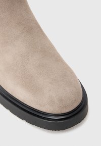 Botte de cheville en daim beige avec un bout rond et une semelle en caoutchouc noir. Présente une texture lisse et un design minimaliste sans éléments décoratifs.