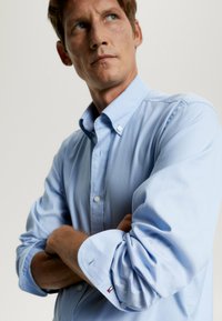 Camicia azzurro chiaro con colletto, maniche lunghe e polsini arrotolati, con un piccolo logo sulla manica. Tessuto liscio.