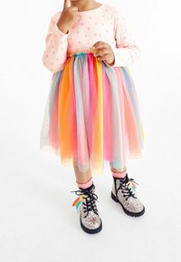 Rosa långärmad klänning med prickar, lager av tyllkjol i regnbågens färger. Glittriga svarta stövlar med regnbågsdetaljer och rosa strumpor.