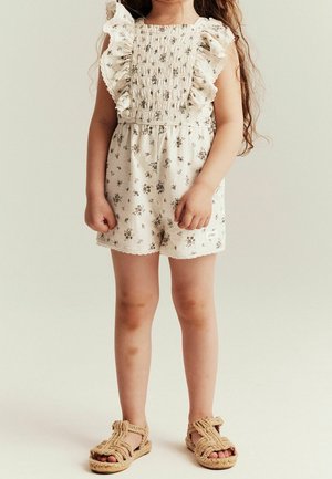 Enfant portant une combinaison crème sans manches à imprimé floral avec des épaules à volants, chaussé de sandales beige tissées, debout devant un fond uni.