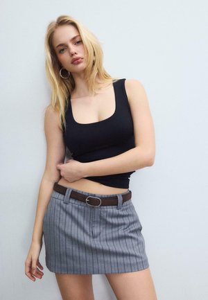 Plavolaska ženska v črni brezrokavnik majici, siva mini krilo z drobnimi črtami in rjavim pasom, ki pozira ob beli steni.