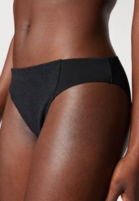 Fekete bikini alsó, lakk részletekkel az egyik oldalon és sima anyaggal a másikon. Magasan szabott dizájn, kényelmes elasztikus derékpánttal.