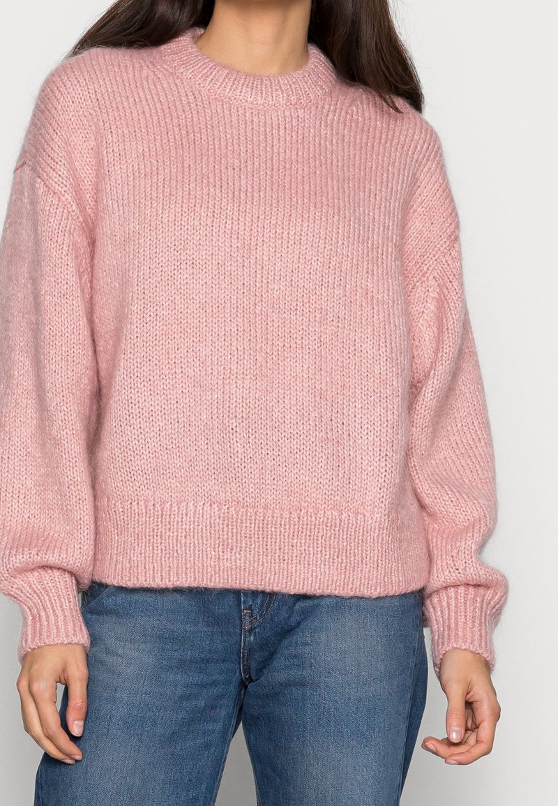 Femme portant un pull rose doux, surdimensionné et en maille, associé à un jean denim bleu moyen sur un fond uni.