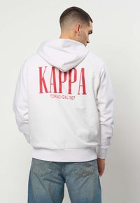 Mees, kes kannab valget kapuutsi punase "KAPPA TORINO-DAL 1967" tekstiga seljal, sinised teksad, näoga eemale ühevärvilise tausta ees.