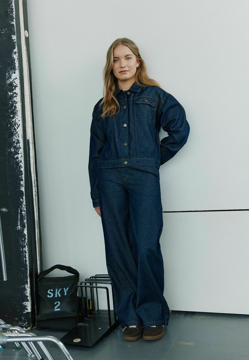 Denim obleka, ki vključuje prilegajočo se jakno in široke hlače v temno modri barvi. Jakna ima zaponke na gumb in tkanina ima gladko teksturo.