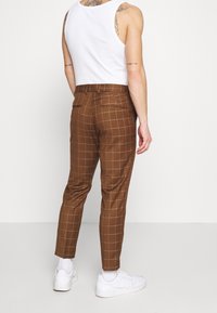 Brown checkered trousers made of fabric, featuring a slim fit, two back pockets, and rolled hems. Worn with a white tank top and white sneakers.