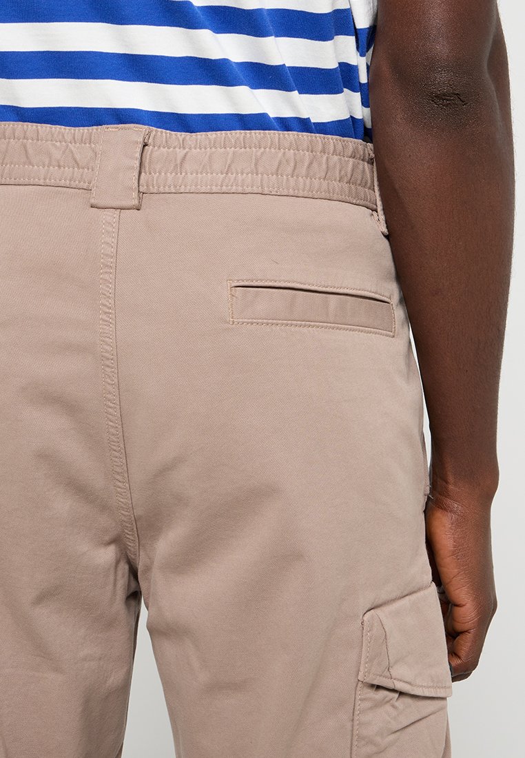 Beige cargobroek met een elastische tailleband, zijzakdetails en een gladde katoenstructuur, gecombineerd met een blauw-wit gestreept bovenstuk.