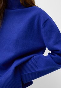 Knit sweater in vibrant blue, featuring a high ribbed collar and wide ribbed cuffs, with a soft texture and relaxed fit.
