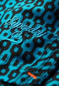 Bañadores en turquesa y negro con un patrón geométrico. El nombre de la marca "Superdry" está bordado en hilo azul.