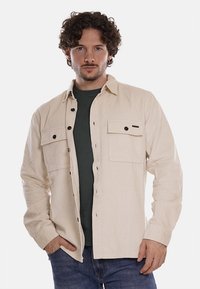 Camicia in corda beige chiaro con chiusura a bottoni, due tasche sul petto e dettagli in bottone neri, indossata sopra una maglietta verde scuro.
