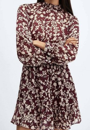 Kobieta ubrana w bordową sukienkę z długim rękawem, ozdobioną kremowymi kwiatowymi wzorami, z falbaną na dole, ze skrzyżowanymi ramionami na klatce piersiowej.