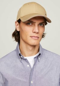 Cappello da baseball in tessuto beige, con visiera strutturata e cuciture a contrasto. Indossato con una camicia azzurro chiaro a maniche lunghe.