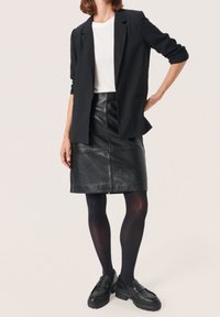 Blazer nero abbinato a una maglietta bianca, gonne a tubino in pelle nera e collant scuri. L'outfit presenta linee eleganti e un design classico.