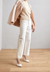 Person som bär en beige kavaj över axlarna, en krämfärgad tröja, vida vita byxor och vita högklackade skor, står på ett trägolv.