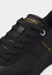 Αθλητικό παπούτσι Black Jack & Jones με υφή δέρματος, μαύρα κορδόνια και χρυσό λογότυπο στο πλευρό και στη γλώσσα, σε λευκό φόντο.