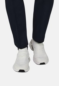 Chaussures de sport blanches en tissu texturé, avec des lacets et une semelle extérieure noire, portées avec un pantalon bleu marine foncé à coupe fuselée.