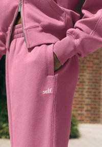 Rúžový set mikiny a nohavíc vyrobený z mäkkého materiálu, s detaily na zips, ribbovanými manžetami a vyšitým logom "self." na vrecku.