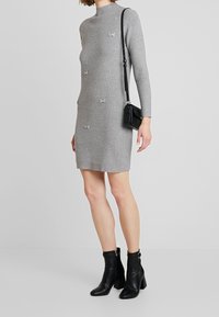 Vestito grigio a coste con maniche lunghe, dotato di fiocchi decorativi e perline. Abbinato a stivaletti neri in pelle fino alla caviglia e a una piccola borsa a tracolla.