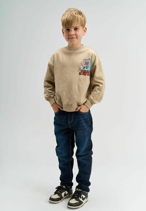 Bežs pulover z barvitim grafičnim besedilom in oblikovanjem na prsih, v kombinaciji s temno modrimi jeans hlačami in črnimi športniki.