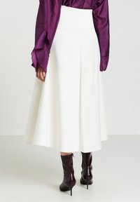 Jupe blanche longue jusqu'au sol avec une taille haute, réalisée en tissu léger. Associée à des bottes à talons hautes jusqu'aux genoux de couleur bordeaux.