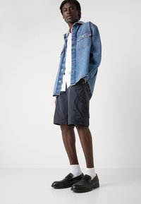 Veste en denim bleu clair, chemise blanche, short marine avec poches latérales et mocassins noirs associés à des chaussettes blanches.