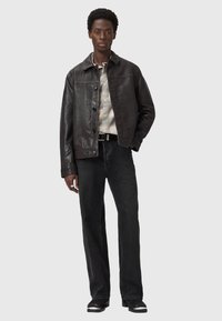 Veste en cuir brun avec fermeture à boutons, deux poches poitrine et un col classique. Portée sur une chemise à motifs, associée à un jean foncé.