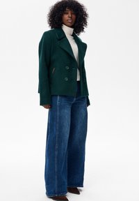 Kobieta z kręconymi włosami ubrana w zielony dwurzędowy płaszcz, biały golf, szerokie niebieskie dżinsy i brązowe czółenka, stojąca.