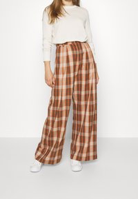 Široké kostkované kalhoty v hnědé, oranžové a béžové barvě s vysokým pasem, zdobené ostrými záhyby a měkkou tkaninou, doplněné bílými botami.