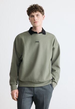 Młody mężczyzna o kręconych włosach, ubrany w oliwkowozieloną bluzę z logo "BOSS" oraz szare spodnie, stojący na jednolitym jasnym tle.