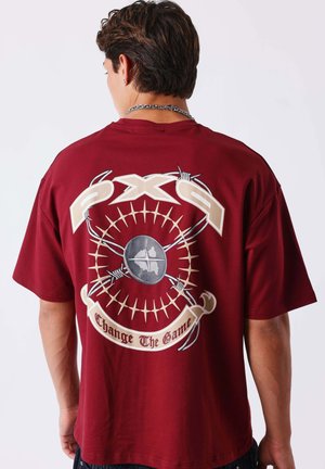 Personne portant une chemise bordeaux avec un grand motif de fil barbelé, un emblème circulaire et le texte "Change The Game" dans le dos.