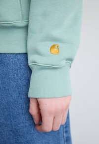 Mintgrüner Pullover mit gerippten Bündchen und einem goldenen gestickten Logo am Ärmel. Kombiniert mit einem Jeansrock.