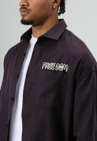 Dikke geruite blouse in bordeauxrood en marineblauw met zwarte knopen; heeft geborduurde tekst "FREE DOM" op het borstzakje; gemaakt van een katoenmix.